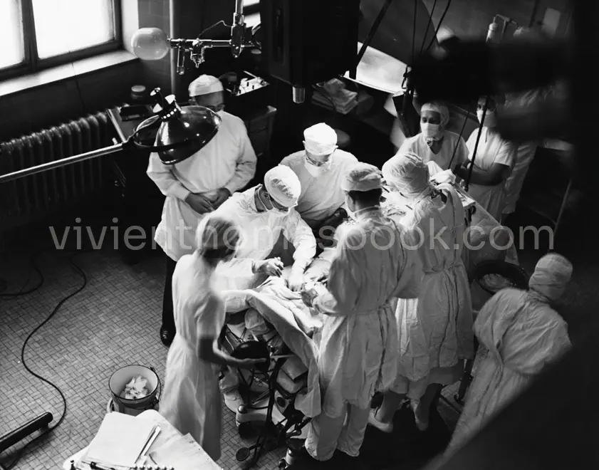 In this 1947 Blue Baby surgery photo, the first released to the press by Johns Hopkins University, Vivien Thomas remained unidentified and uncredited. He was also posed ungloved and was partially hidden behind the standing surgical lighthead as he advised Alfred Blalock through the surgery.